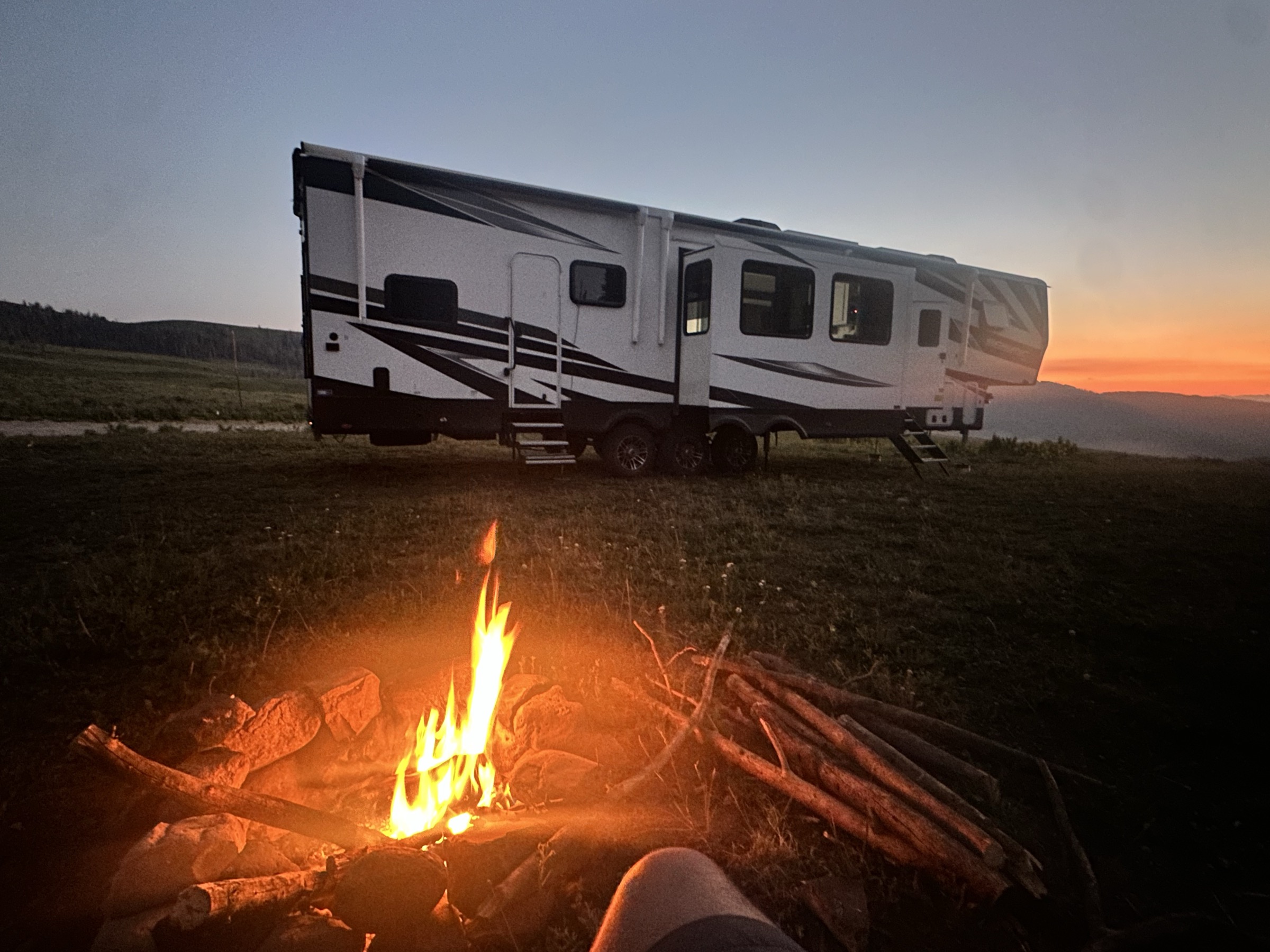 A 5th-wheel parked at a high-desert dispersed site at sunset with a small campfire
